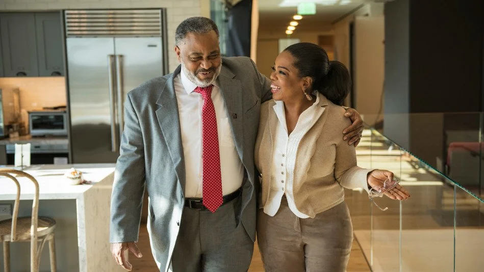 A man and woman are walking together in a modern kitchen or office space, smiling and engaging in conversation. The man has gray hair and a beard, wearing a gray suit with a red tie, and the woman has dark hair tied back, wearing a beige blazer and white shirt.