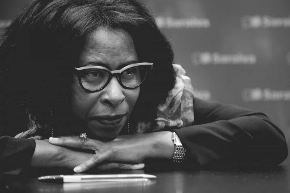 A woman wearing glasses rests her head on her crossed arms on a table, with a pen and a watch visible on her wrist.