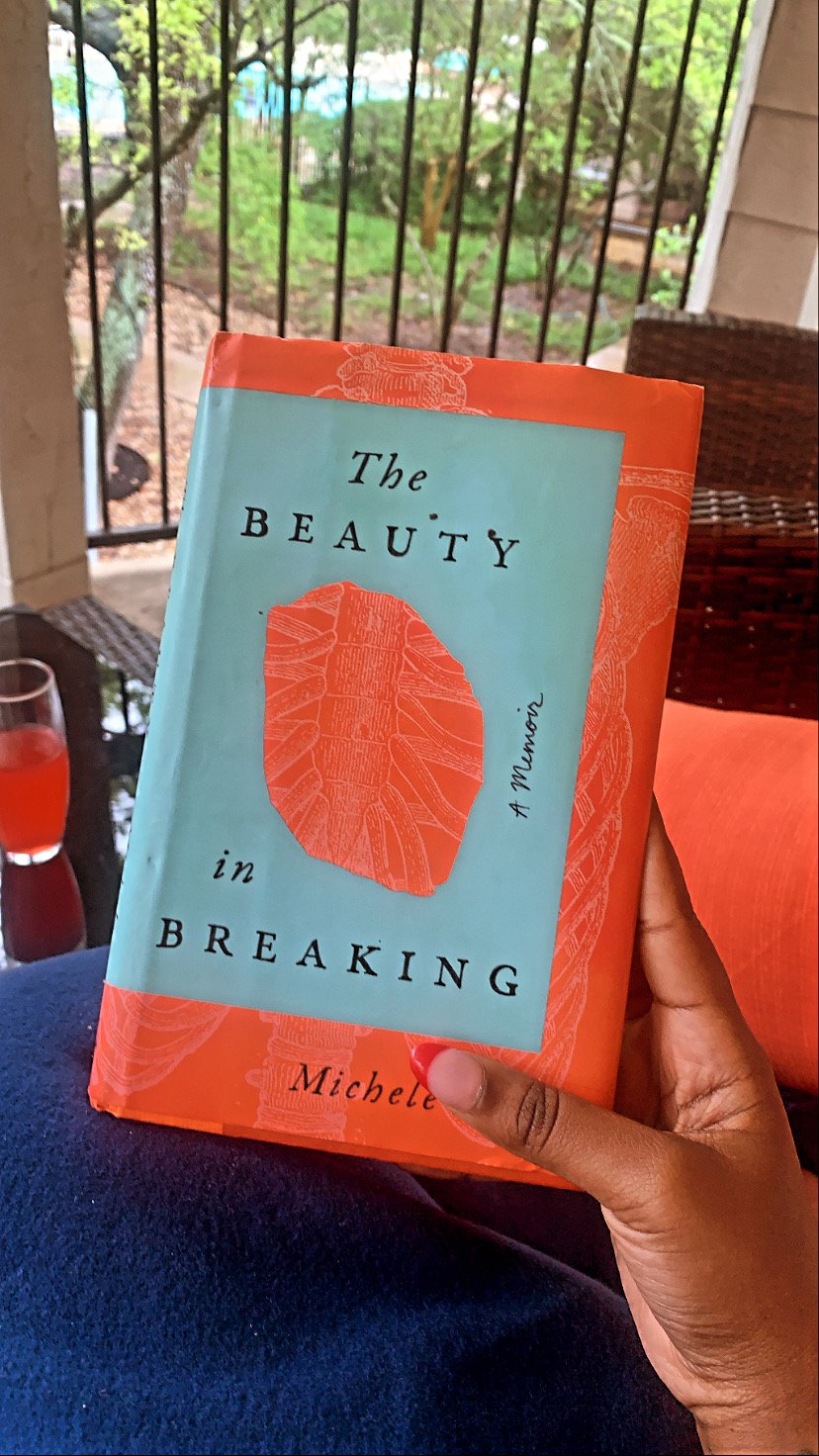 A person holding a book titled "The Beauty in Breaking" by Michele in front of a screened porch with a view of trees and greenery outside.