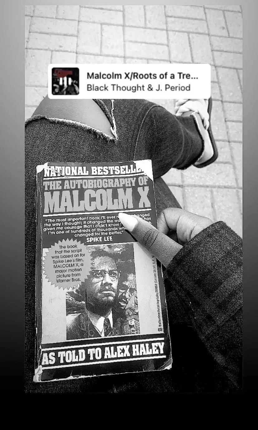 A black-and-white photo of a person sitting outside on a sidewalk, holding a newspaper about Malcolm X. The person is wearing flip-flops, ripped jeans, and a dark sweatshirt. The background shows pavement tiles.