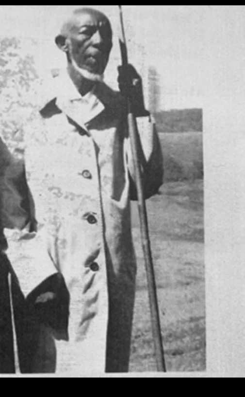 An elderly man wearing a military uniform and glasses, holding a cane and standing outdoors.