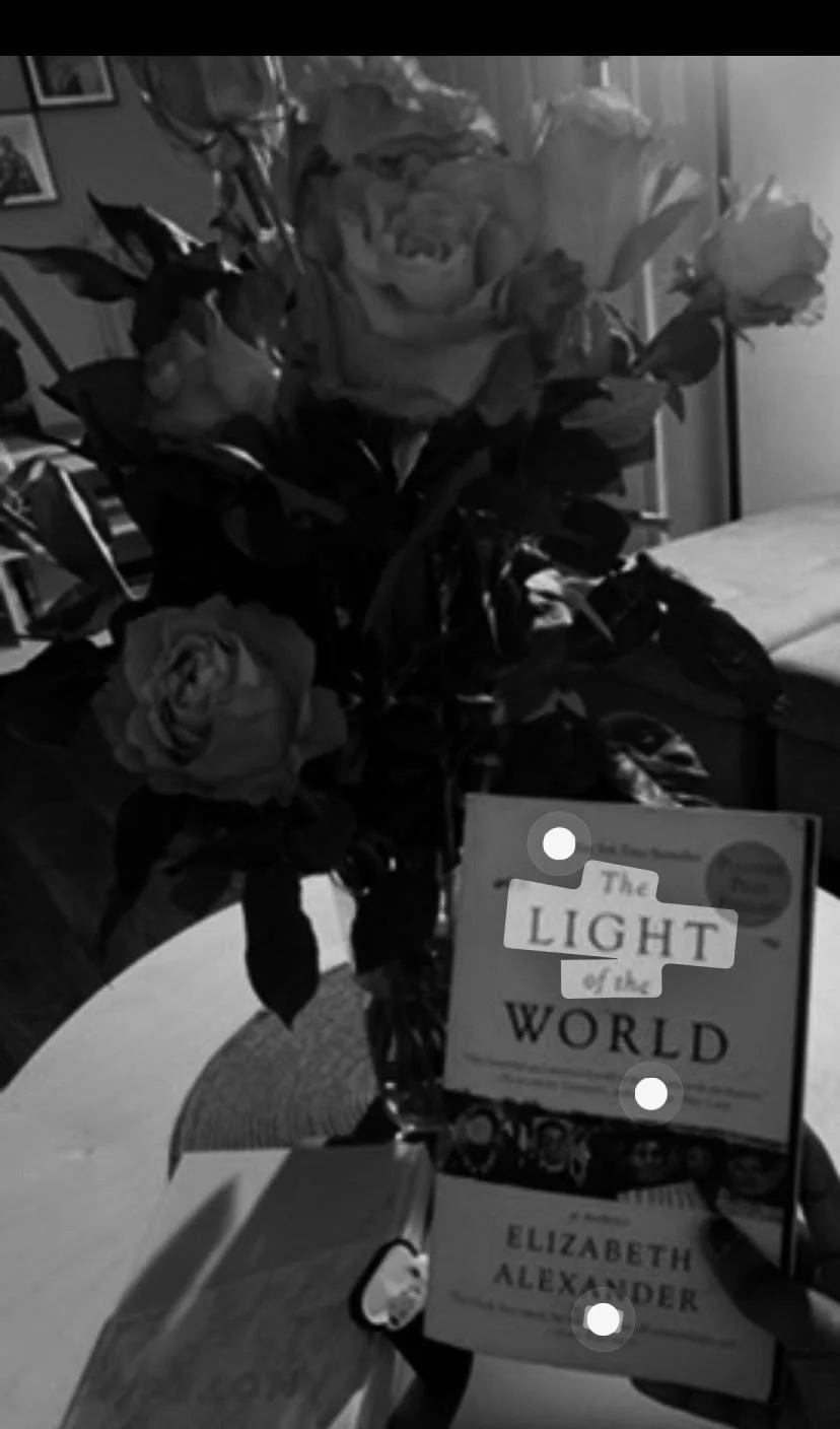 A bouquet of roses with two books, "The Light of the World" by Elizabeth Alexander and another book, on a wooden table in a living room.