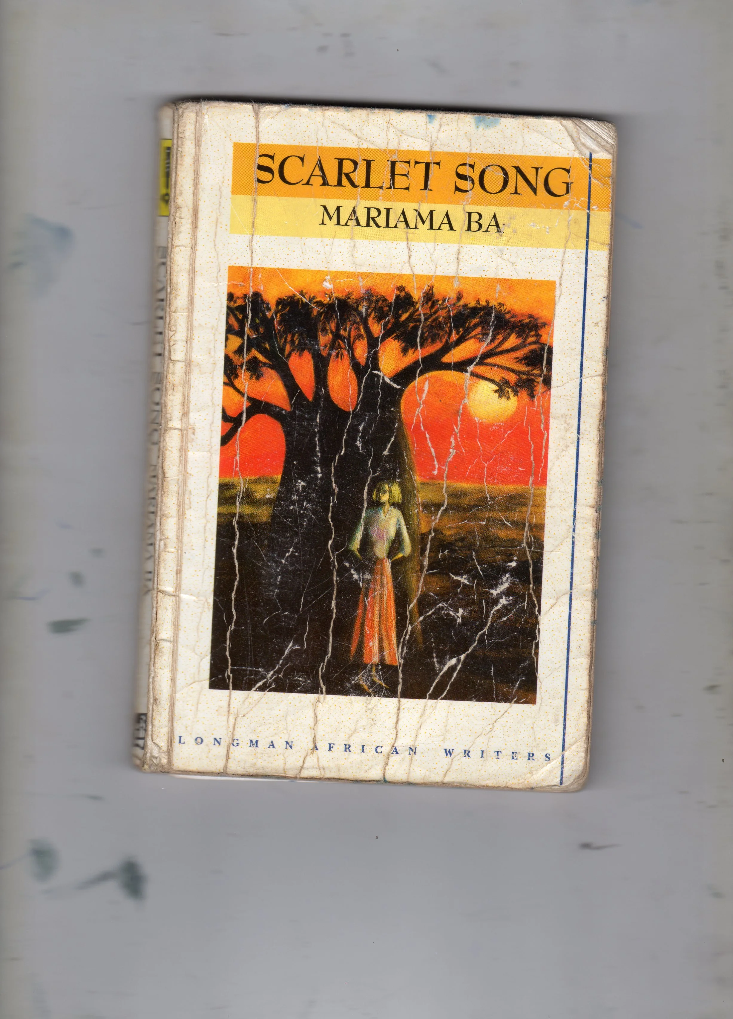 Well-worn book titled "Scarlet Song" by Mariama Bâ, featuring an illustration of a woman standing in front of a large tree during sunset.