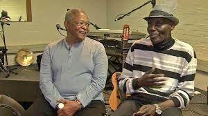 Two people sitting and talking in a room, smiling, with musical instruments and equipment in the background.