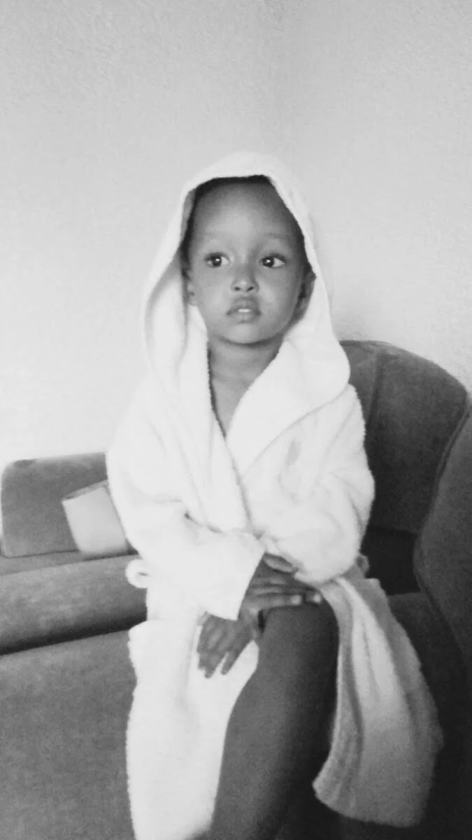 A young child sitting on a couch, wearing a hooded towel, with a neutral expression.