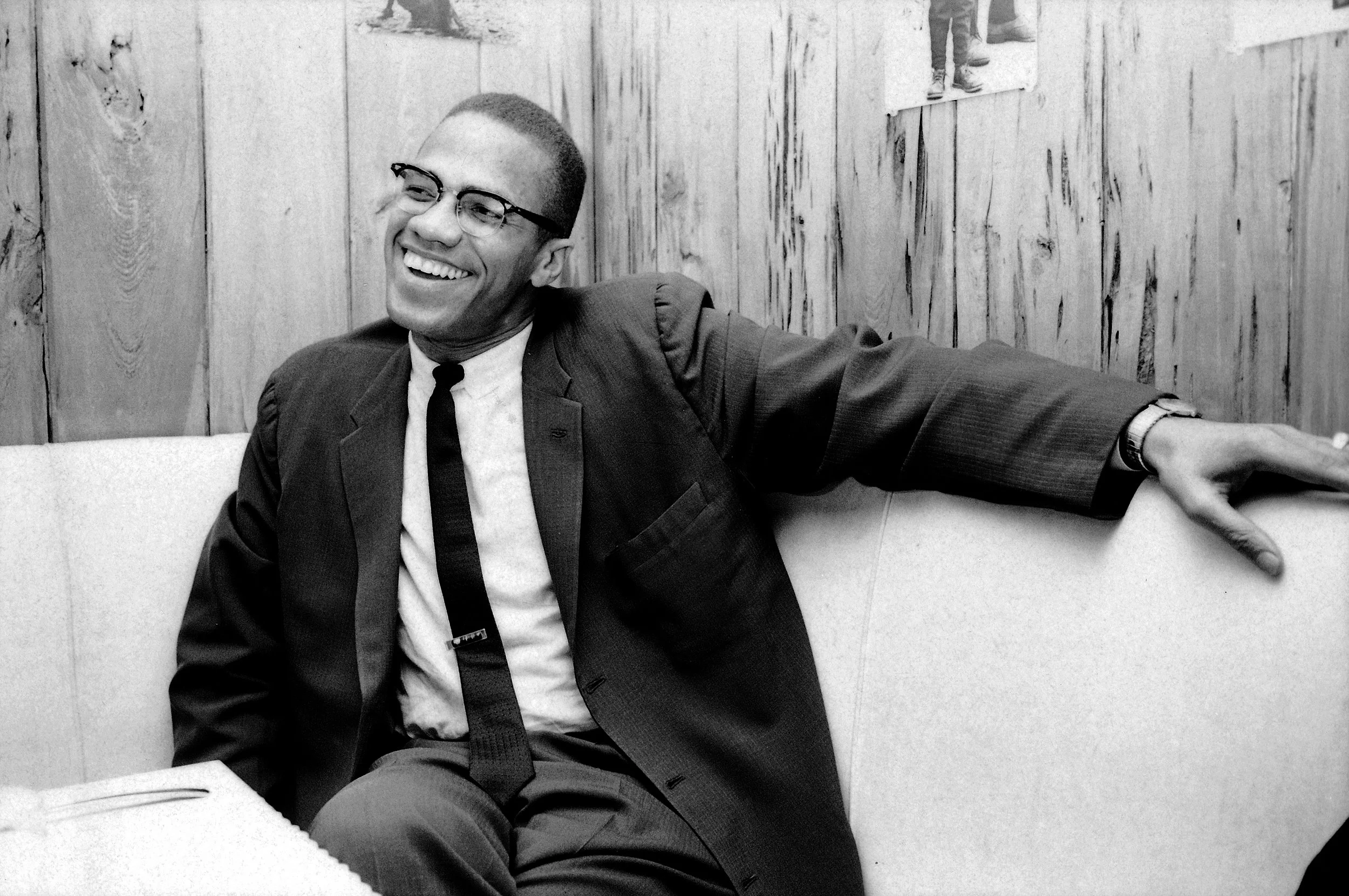 A smiling man in a suit and tie with glasses, sitting on a booth seat in a wooden-paneled room, with his arm extended along the back of the seat.