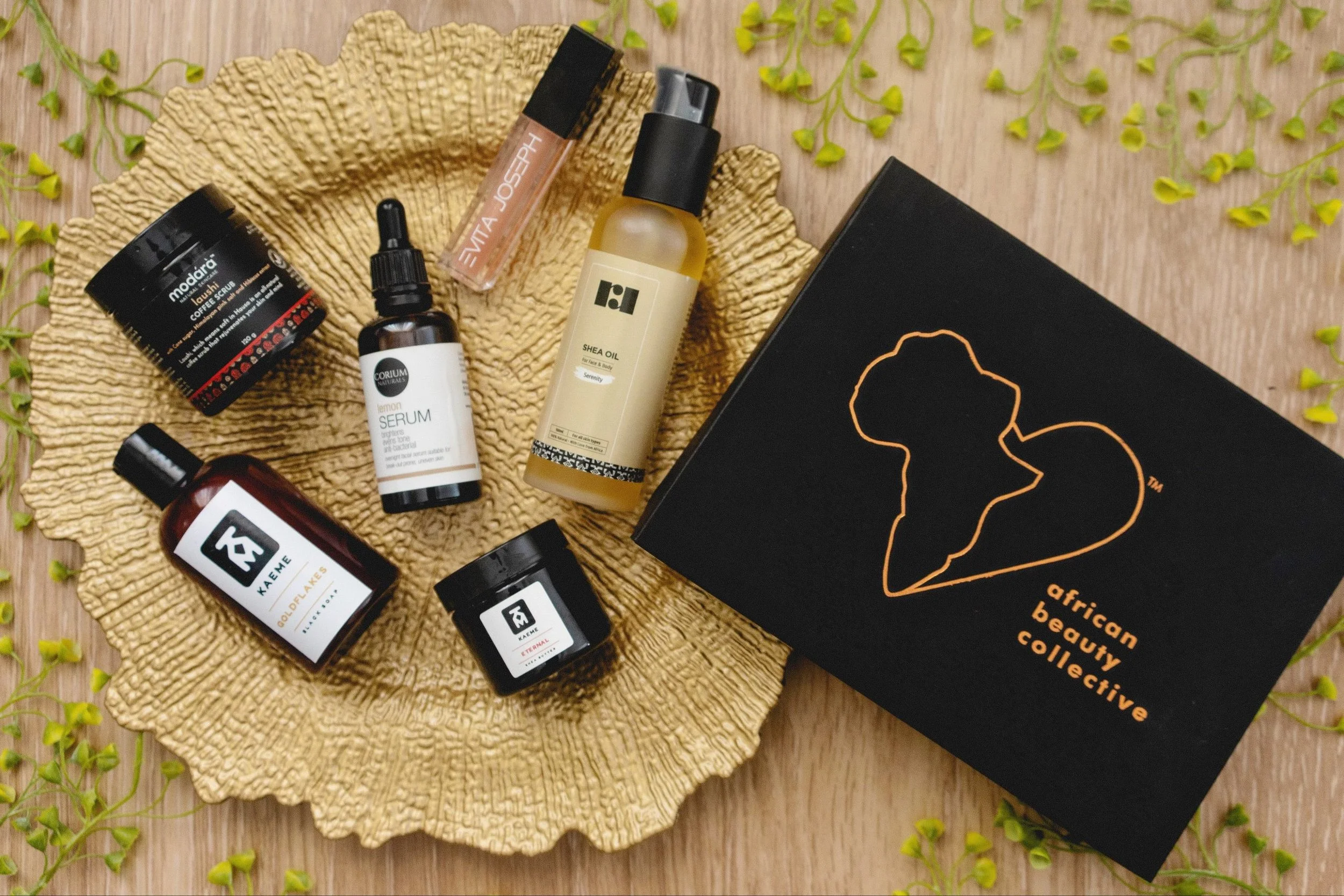 Various skincare and wellness products arranged on a yellow textured plate, including creams, serums, oils, and a lip gloss, next to a black box labeled 'African Beauty Collective' with a heart-shaped logo.