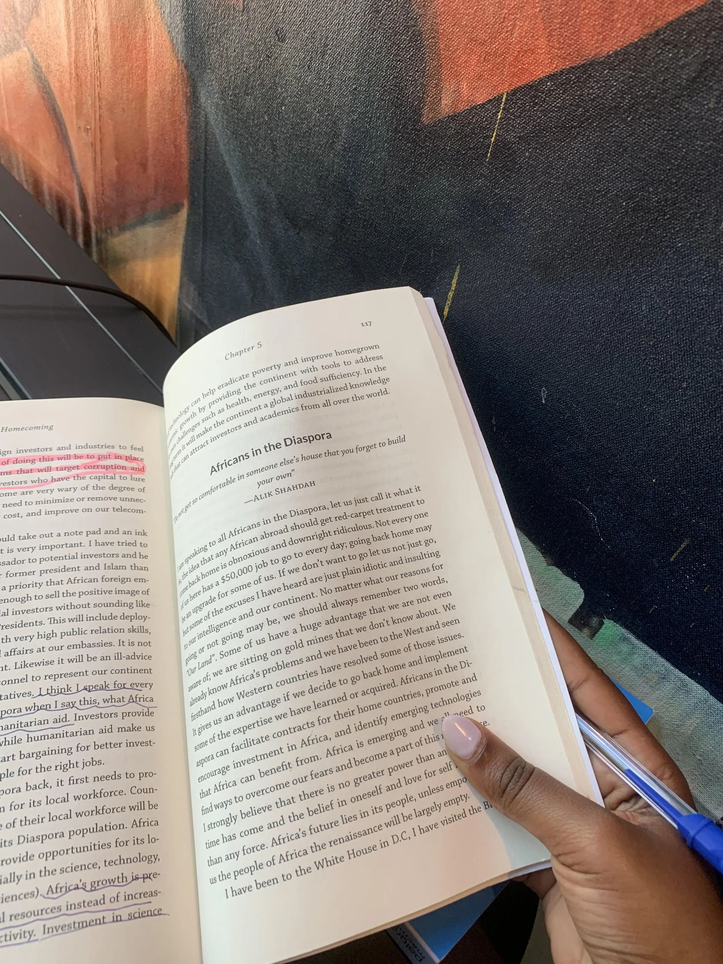 Open book with chapter 5 titled 'Africans in the Diaspora' being held in a person's left hand with a blue pen in the right hand, against a background of a colorful textured fabric and a laptop.
