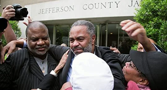 A group of people, including men and women, are gathered outside the Jefferson County jail. They appear to be comforting or consoling a man at the center. One person is taking a photo with a camera.