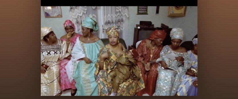 A group of women in traditional colorful African attire seated indoors, some wearing head wraps, engaged in conversation or looking at their phones.