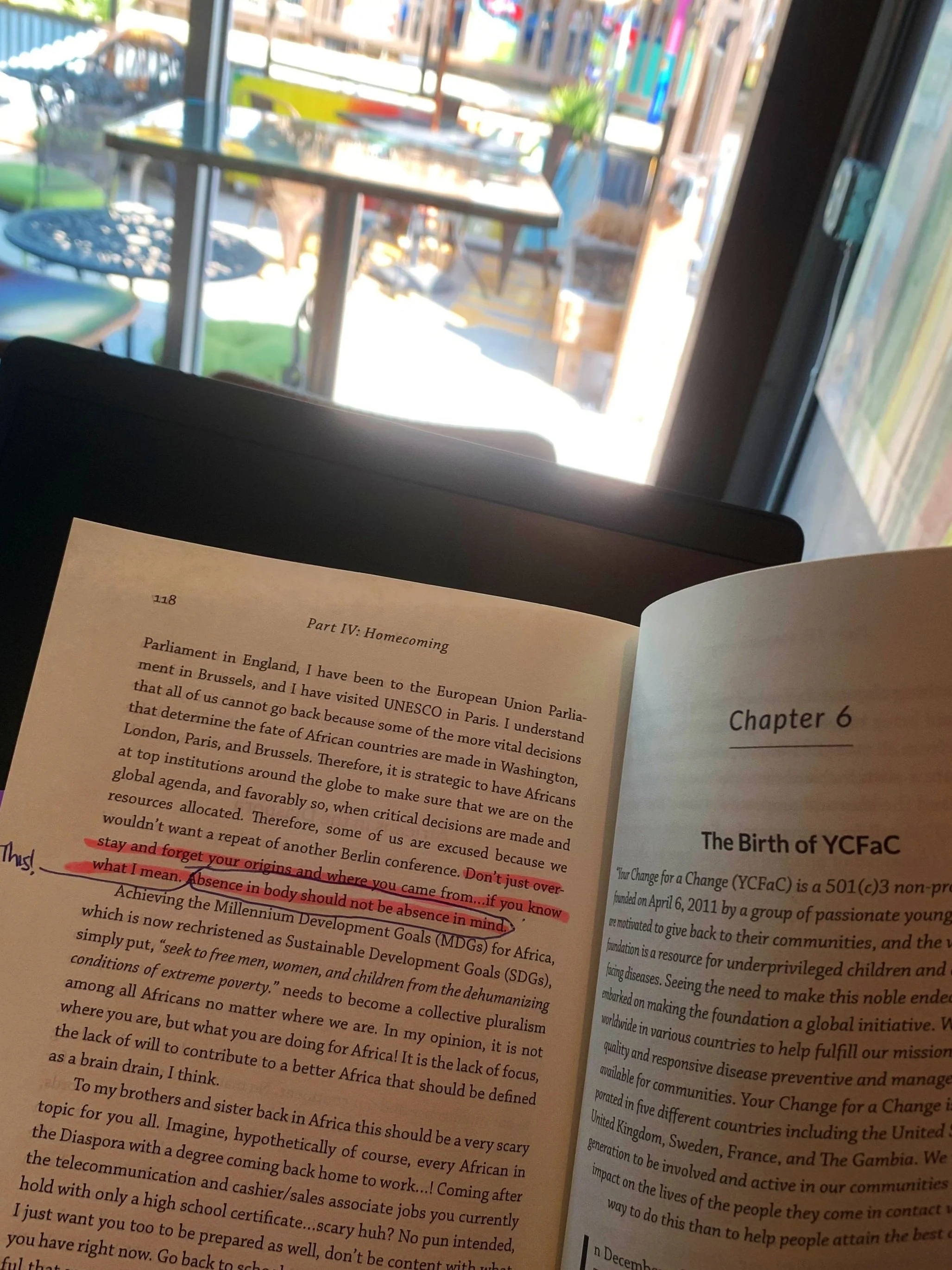 Open book with highlighted text, held in front of a window in a cafe or restaurant. Outside, there are tables, chairs, and colorful decor.