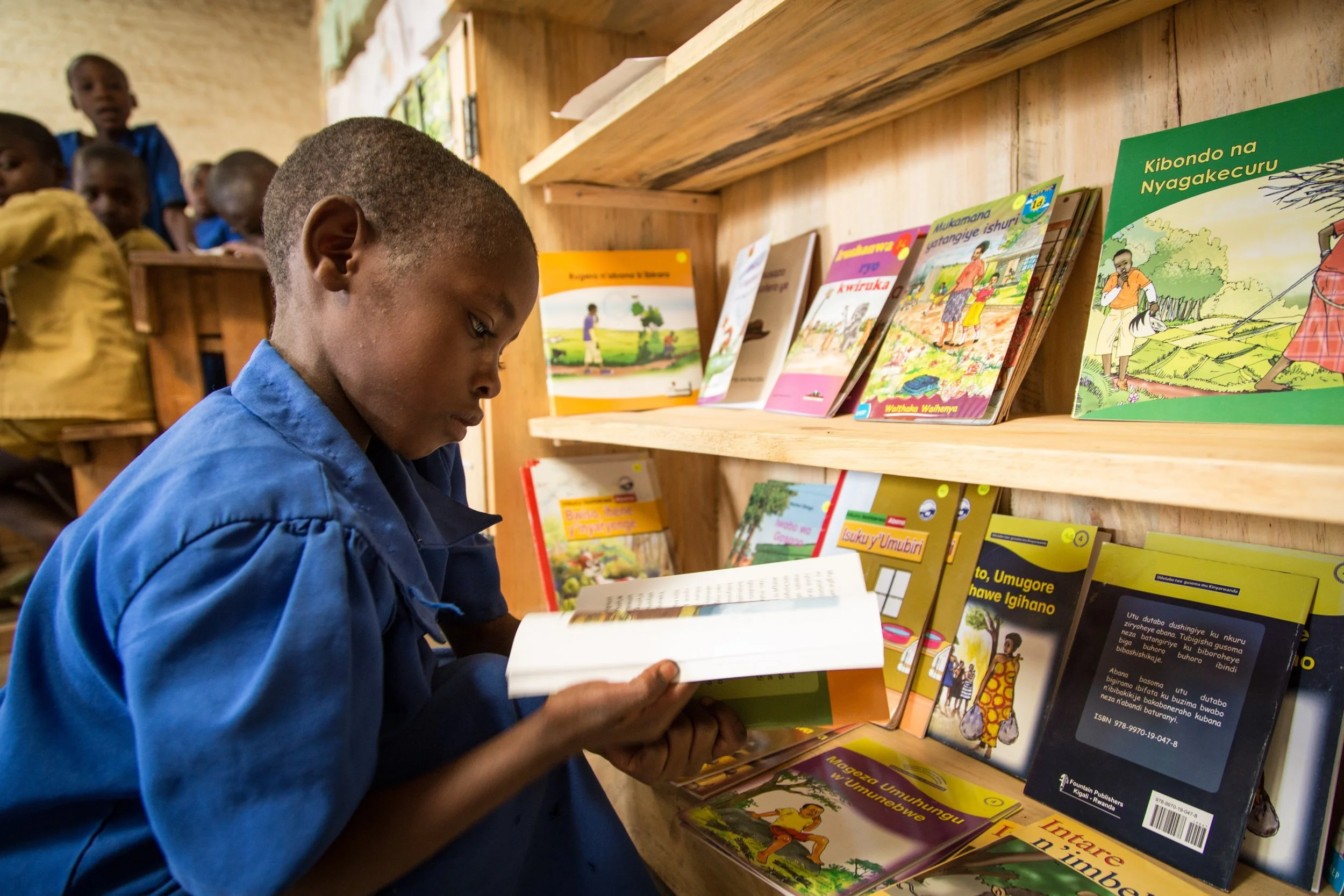 A young boy in a blue school uniform reading a book in a classroom or library, with other children in uniforms sitting in the background and colorful books on a shelf.
