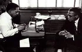 Two men sitting at a table in a restaurant or cafe, engaged in conversation. One man is on the left, and the other on the right, with cups and a napkin dispenser on the table.