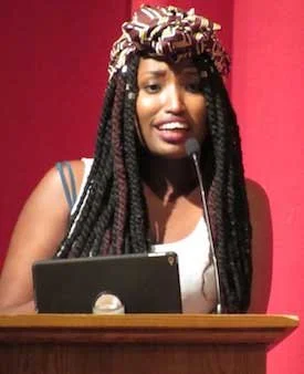 Woman with basket-weave styled braids and a headwrap speaking at a podium