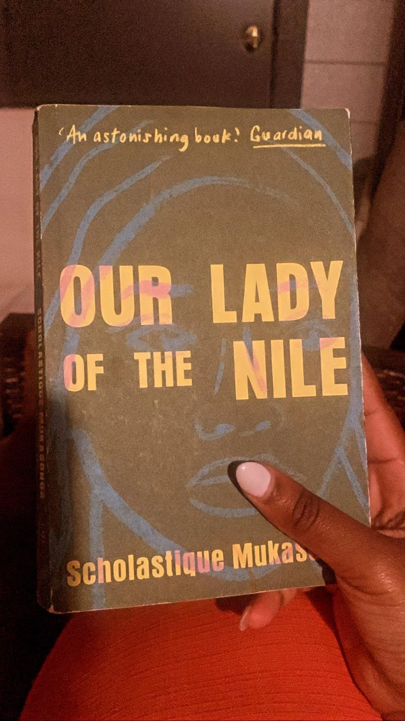 A book titled 'Our Lady of the Nile' by Scholastique Mukasonga, held by a person with dark skin. The cover is gray with pink and yellow lettering and has a partial line drawing of a face in the background.