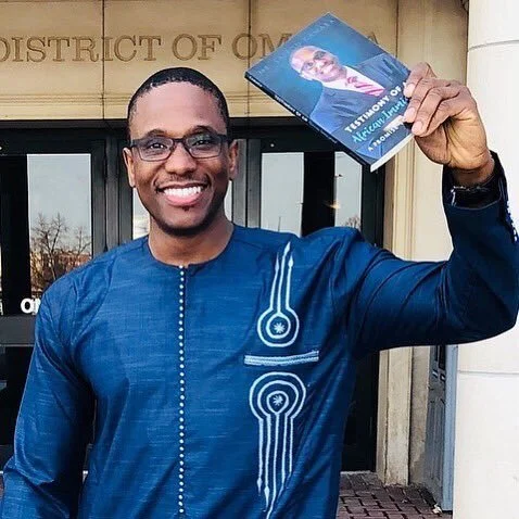 A smiling man in glasses and a blue traditional African outfit holding a book outside a building with a sign that reads 'District of...'