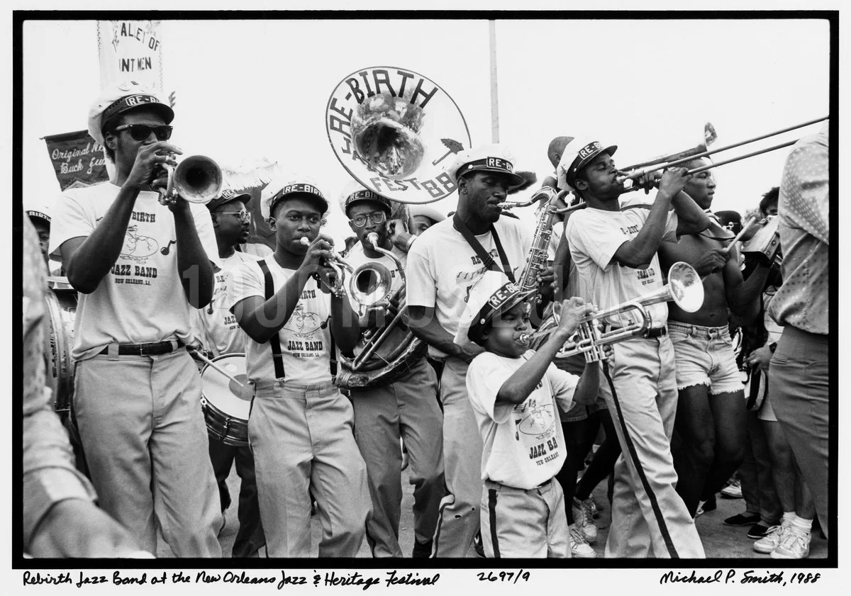 A black-and-white photo of a young jazz band performing at an outdoor festival. The band members are playing various instruments like trumpets, saxophones, and drums. They are wearing T-shirts and hats that read 'Rebirth Jazz Band.' In the background, there is a banner for the New Orleans Jazz & Heritage Festival.