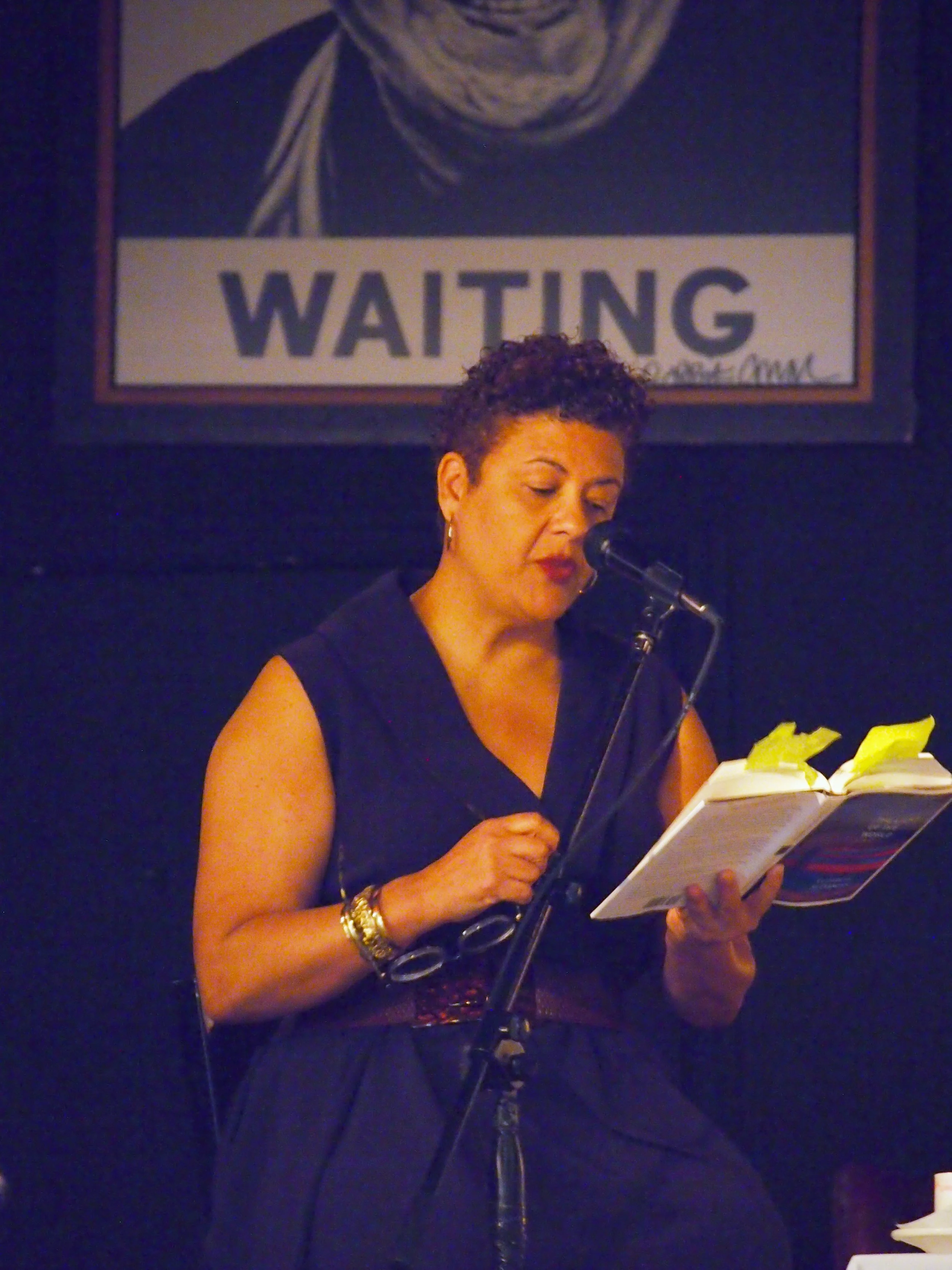A woman reading from a book and speaking into a microphone in front of a sign that says "WAITING."