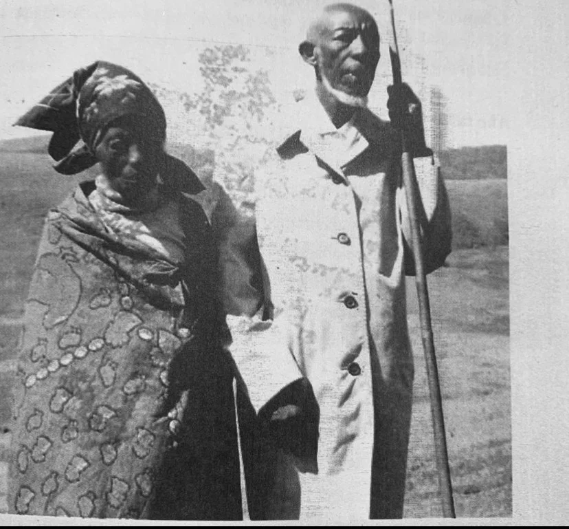 A black and white photo of an elderly man holding a cane and a rifle, standing next to an elderly woman wearing a patterned headscarf and traditional cloth, outdoors.