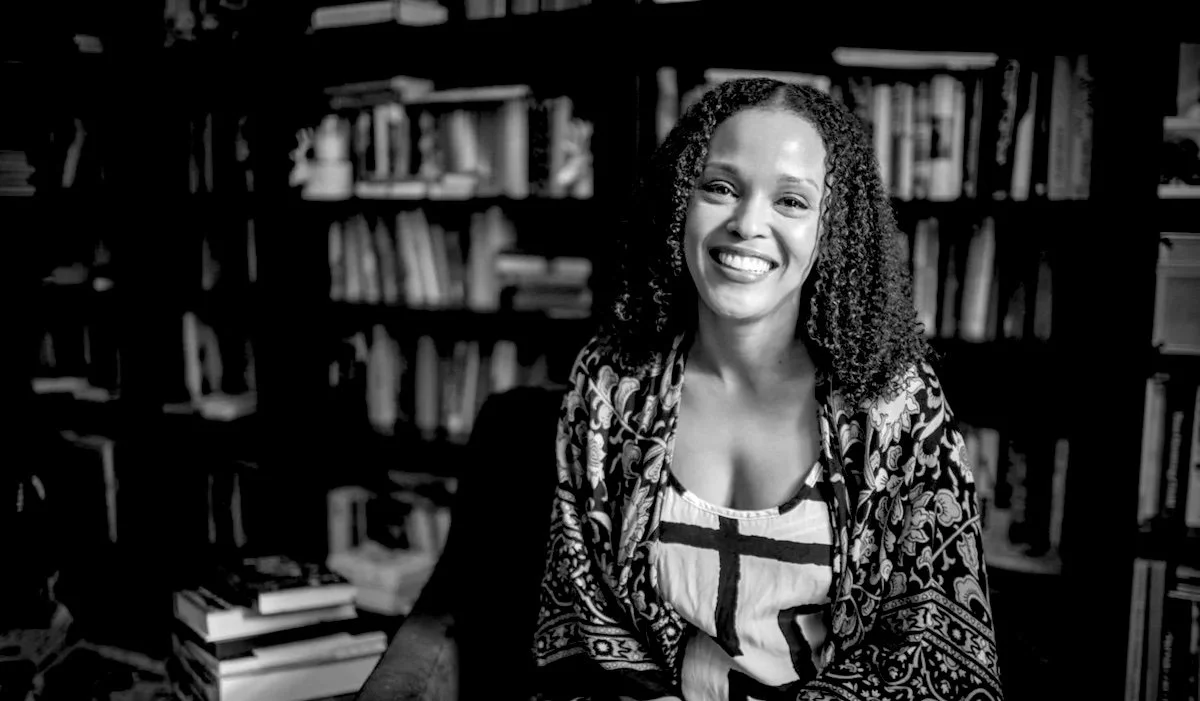 A smiling woman with curly hair sitting in a library with bookshelves in the background.