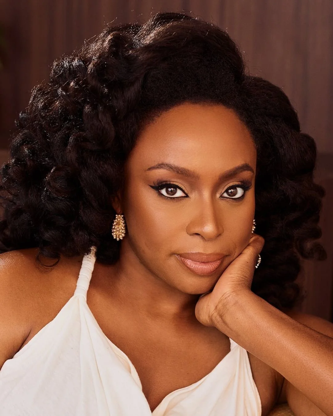 Close-up portrait of a woman with dark curly hair, wearing gold earrings, and a white sleeveless top, resting her face on her hand.