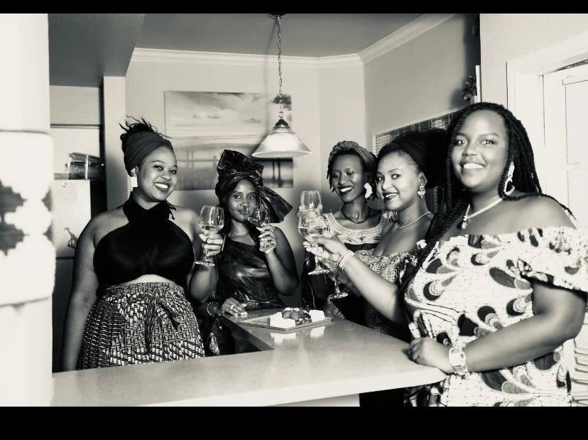 Five women in stylish outfits celebrating with wine glasses in a home kitchen setting.