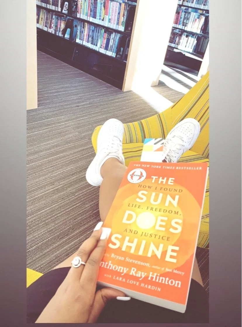A person relaxing in a library or bookstore, sitting on a yellow and green striped chair, holding a copy of the book 'The Sun Does Shine' by Anthony Ray Hinton. They are wearing white sneakers and white nail polish.