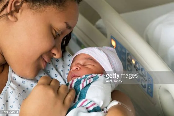 A woman in a hospital bed holding a newborn baby wrapped in a blanket and wearing a hospital hat, looking at the baby lovingly.