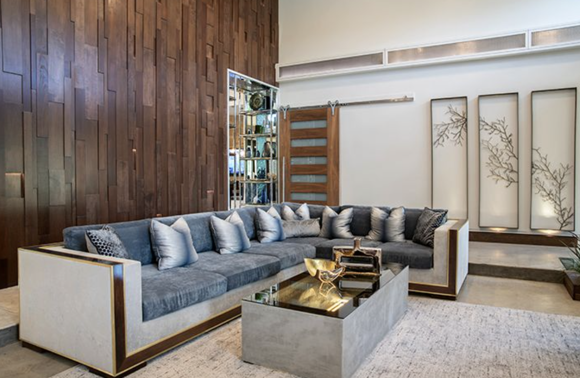 Modern living room with gray sectional sofa, decorative pillows, wooden accent wall, sliding barn door, and three vertical art panels with branch designs, beige carpet, and a coffee table with decorative bowls.