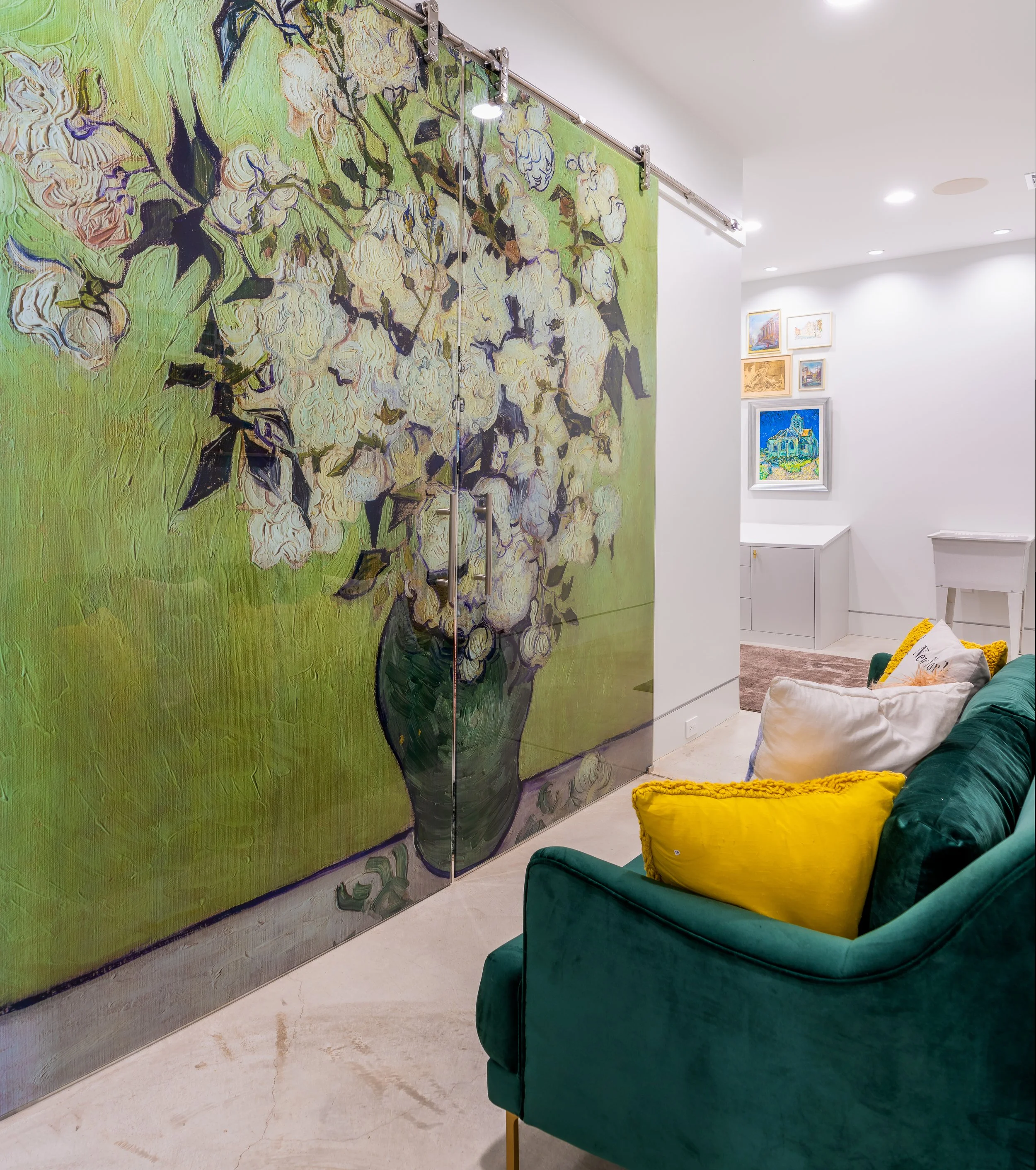 Interior view of an art gallery or living room featuring a large floral painting on sliding doors, a green velvet sofa with yellow and white pillows, and framed artwork on the white walls.