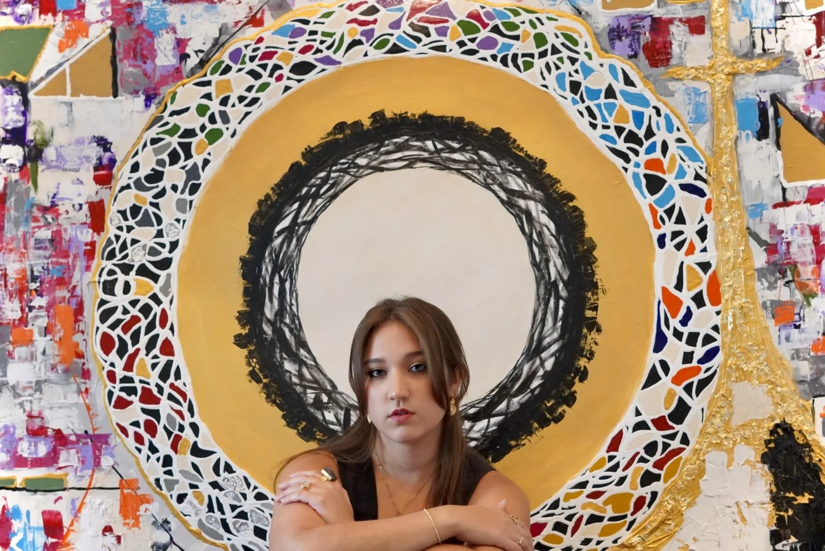 A young woman with long brown hair wearing a black sleeveless top, sitting in front of a vibrant, abstract mural with geometric designs in black, white, yellow, and multicolored sections.
