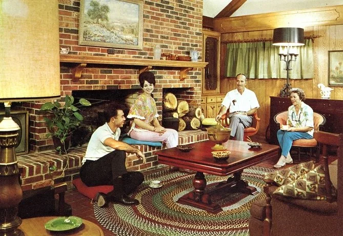 A family sitting in a living room with a brick fireplace, wooden paneling, and a patterned rug. Two women and two men are seated and kneeling around a wooden coffee table.