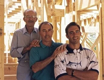 Three men standing together inside a wooden building under construction, smiling at the camera.