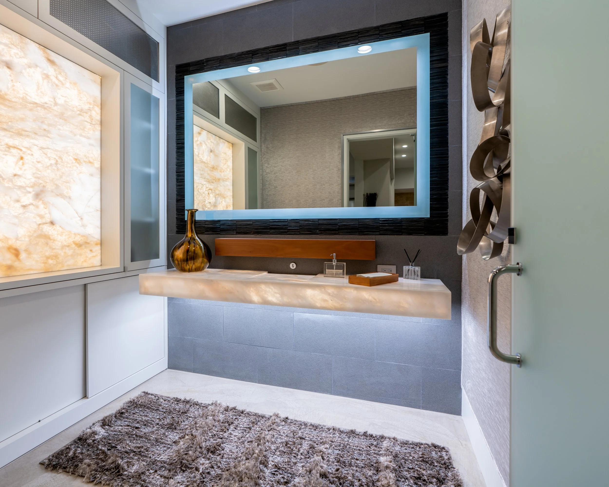 Modern bathroom with a large illuminated mirror, a floating marble vanity with a decorative vase, and abstract metal sculptures on the wall.