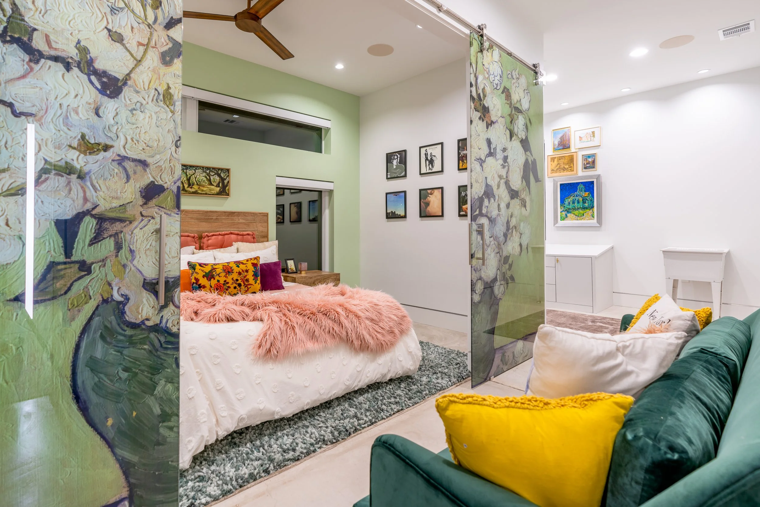 A colorful bedroom with a white bed, pink faux-fur blanket, and multiple pillows, separated by semi-transparent floral glass partitions, and adjacent seating area with a green sofa and various pillows, decorated with framed wall art and a ceiling fan.
