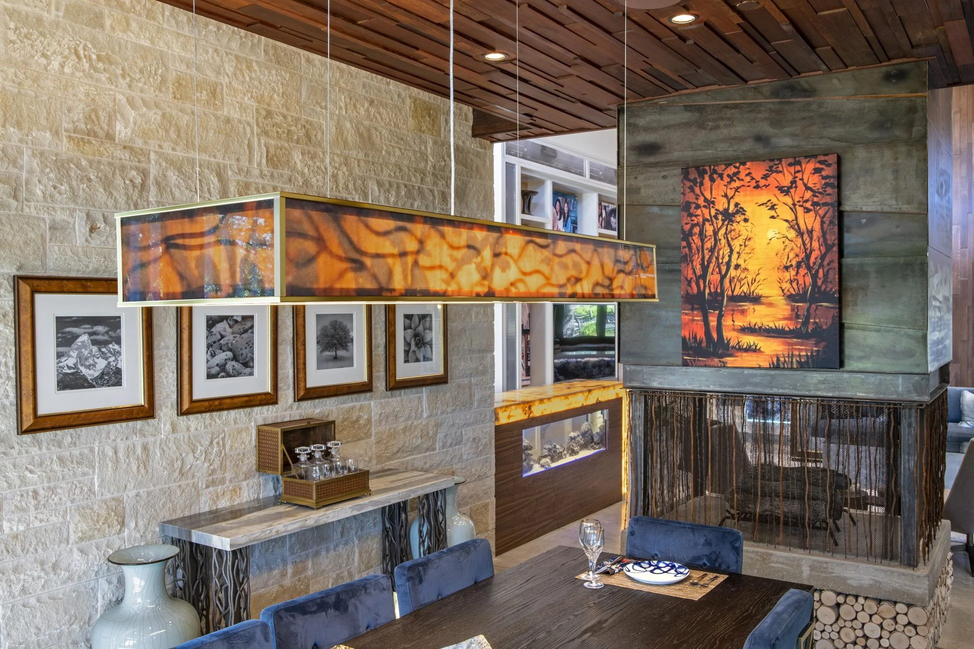 Interior dining room with a dark wood table, blue chairs, and wall art featuring a sunset scene with trees. Modern lighting fixtures hang from the ceiling, and the walls have textured stone and wood paneling.