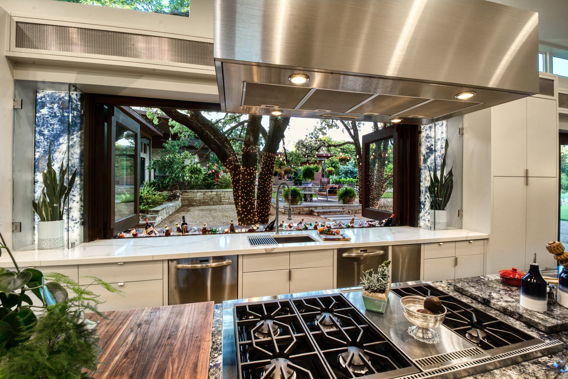 Modern kitchen with a large open window looking out to a backyard with trees and outdoor furniture, decorated with string lights.