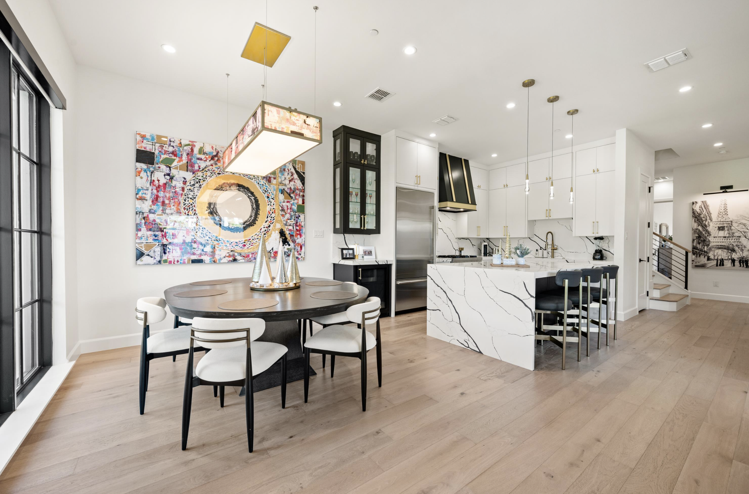 Modern kitchen and dining area with black round table and white chairs, abstract artwork on wall, white cabinets with gold handles, marble island with black bar stools, wood flooring, and a staircase in the background.