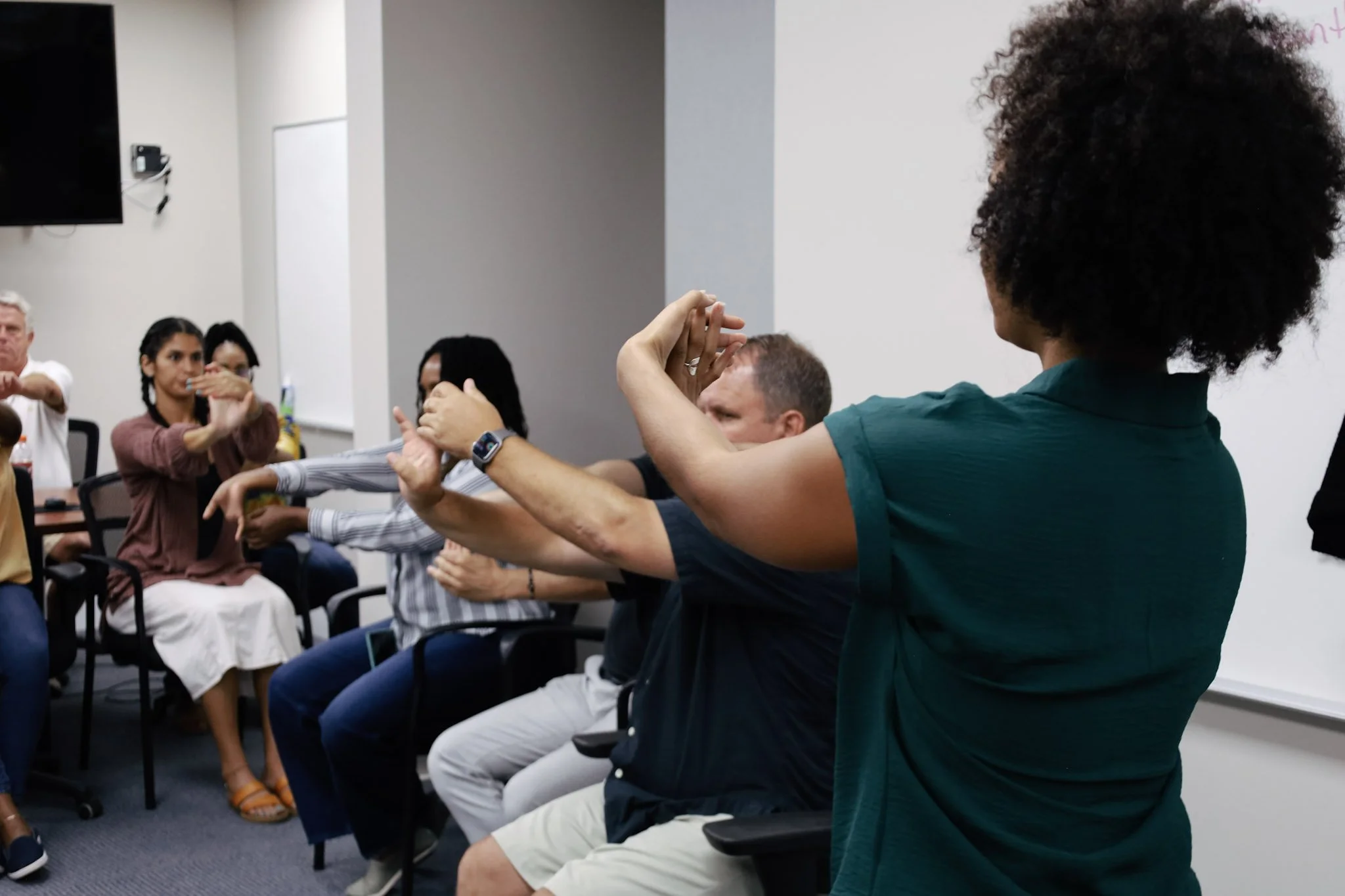 A diverse group of people participating in a seated group activity or workshop, with one woman standing and leading the activity.