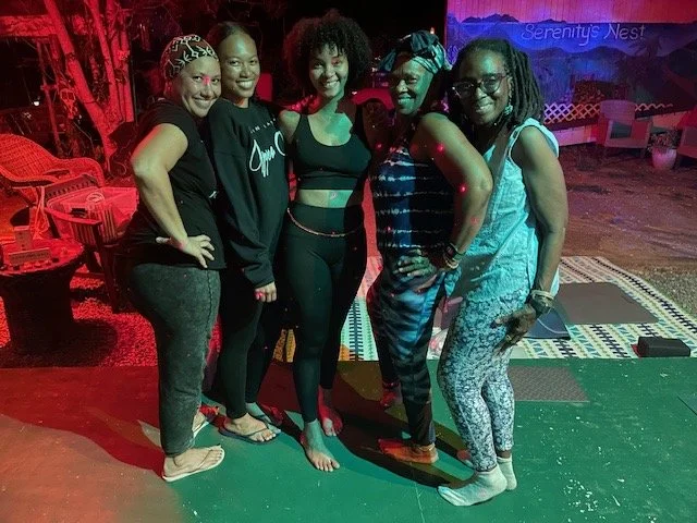 Five women standing together in a dimly lit room, smiling, with colorful lighting and patterned flooring in the background.