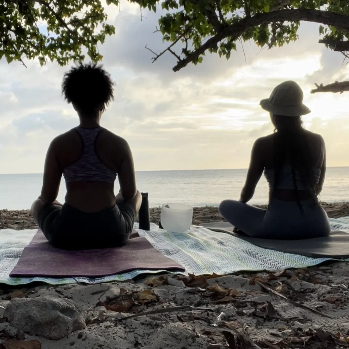 Two women sitting cross-legged on mats on a beach, practicing yoga or meditation during sunset, with the ocean in the background and overhanging trees.