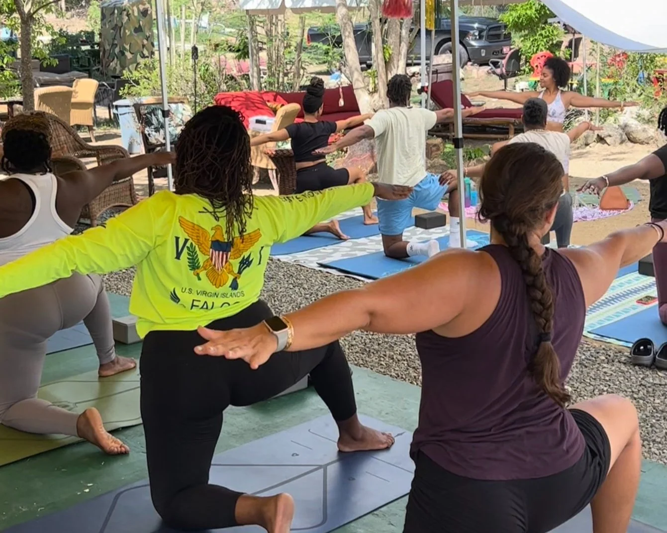 Group of people practicing yoga outdoors, performing Warrior pose on mats under a canopy. Several participants are extending their arms and balancing on one leg amid lush greenery.