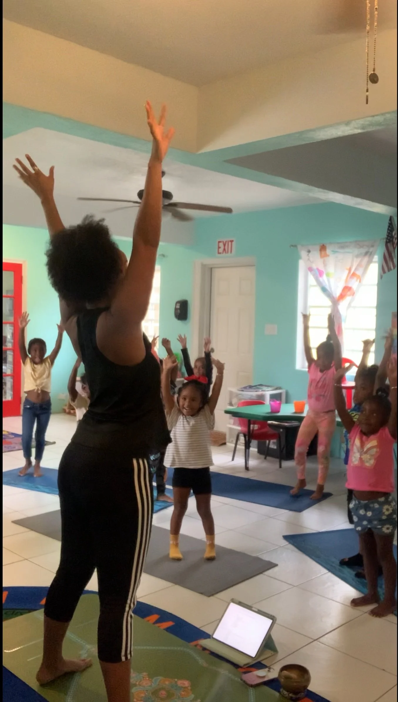 A group of children participating in a yoga or exercise class led by an instructor, standing on mats with their arms raised inside a colorful room.