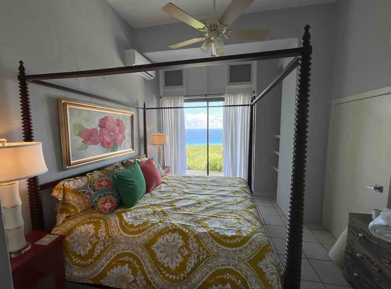A bedroom with a four-poster bed, decorated with patterned yellow and white bedding, and colorful pillows. There is a large painting of pink flowers above the bed. The room has white walls, a ceiling fan, and a large window with ocean view, white curtains, and two small vents near the ceiling.