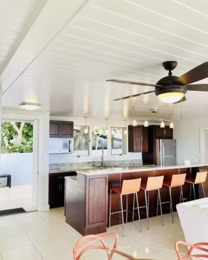 Modern kitchen with dark wood cabinets, a marble island, and four beige bar stools. Ceiling fan and pendant lights over the island. Large windows with outdoor greenery.