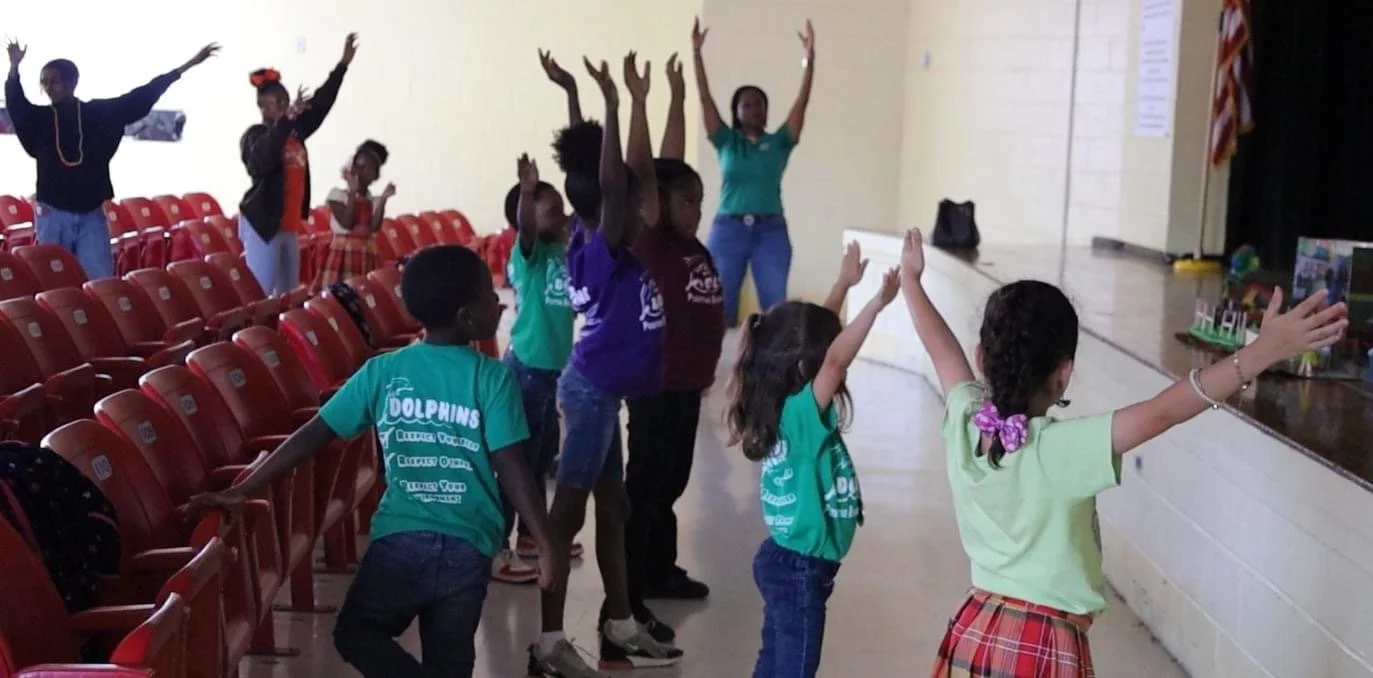Children and adults participating in a group activity on a stage and in an auditorium with red chairs.