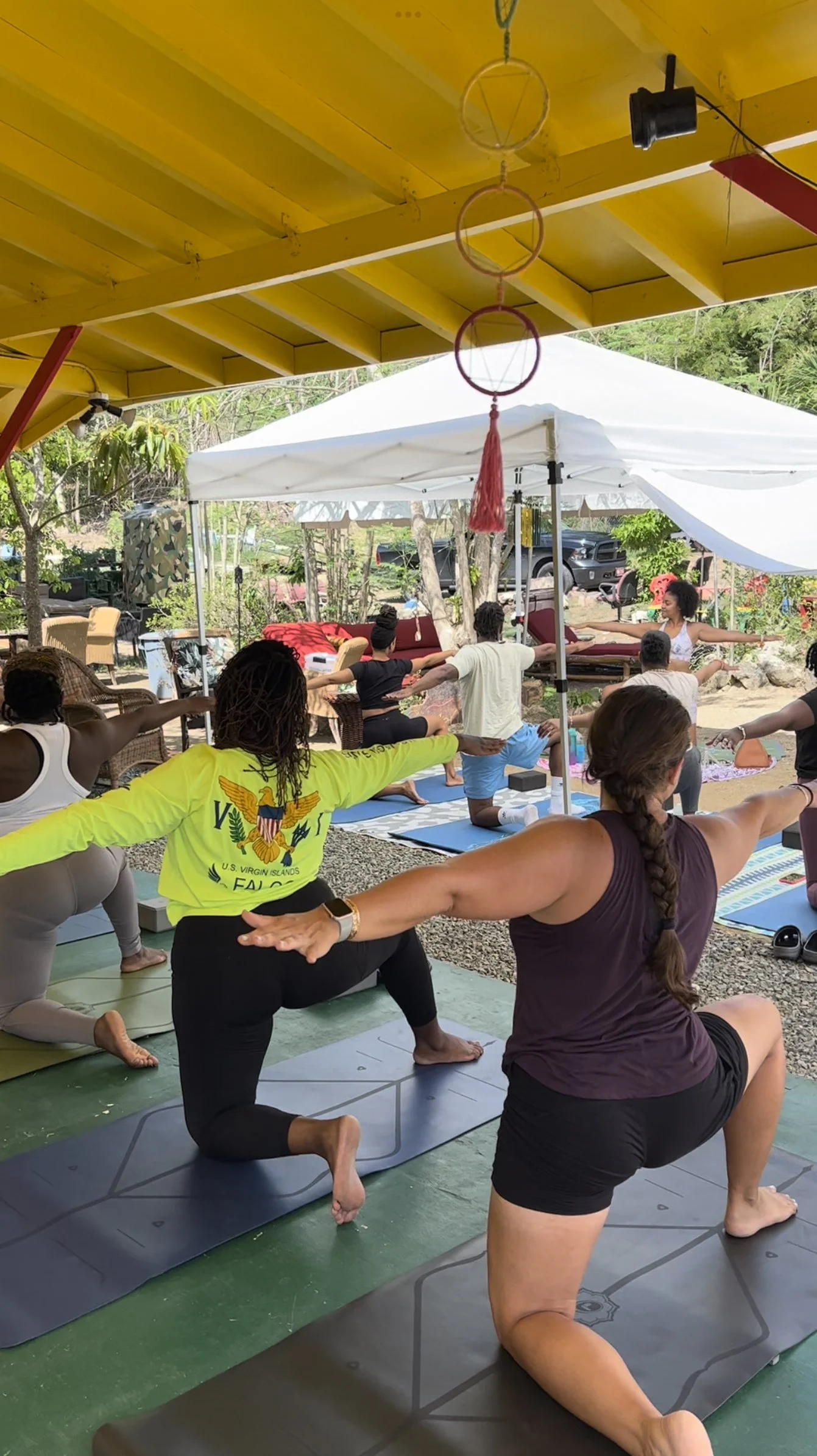 Group of people practicing yoga outdoors under a yellow roof and white canopy on yoga mats, with trees and parked cars in the background.