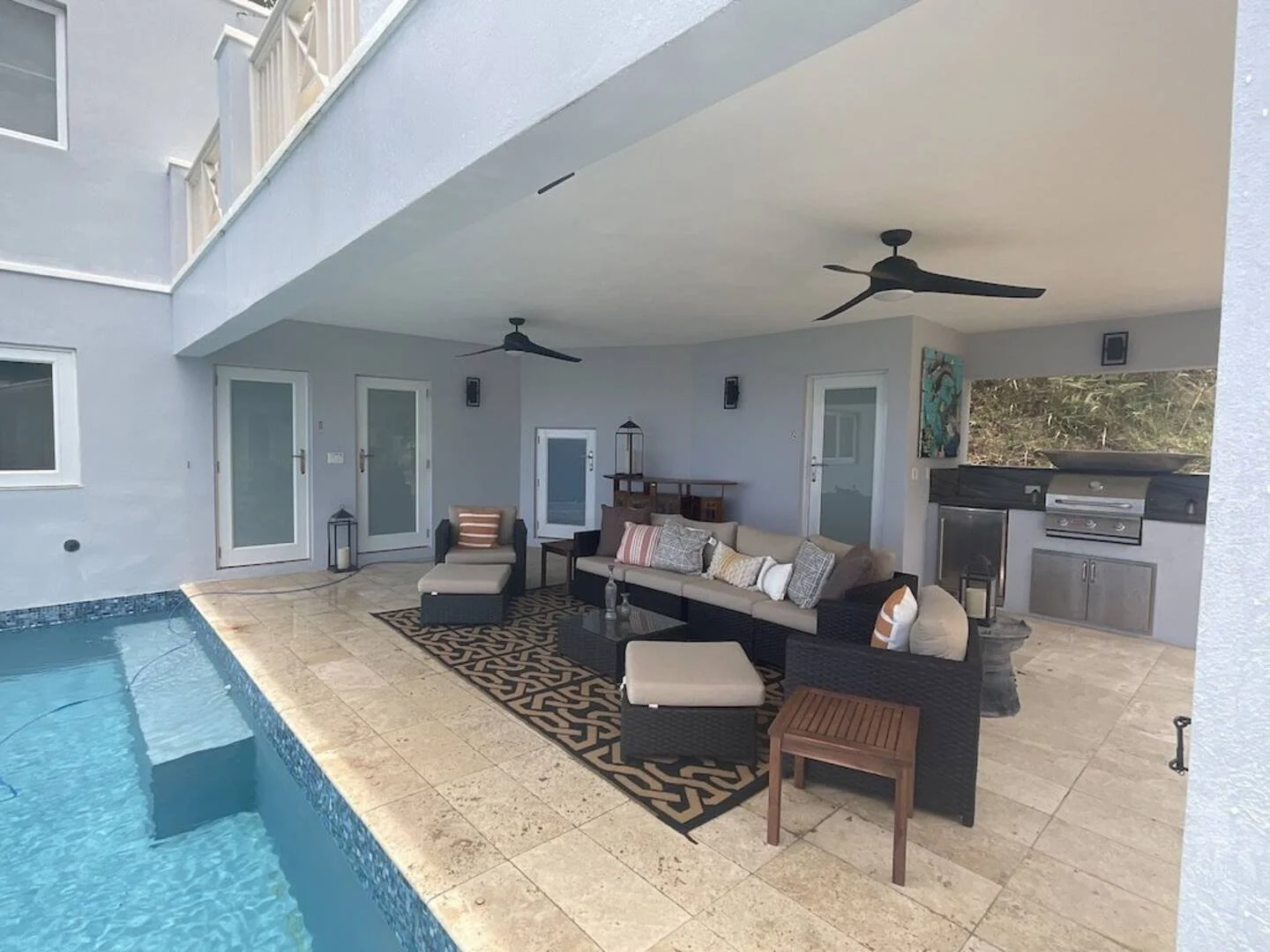 Covered patio area with pool, outdoor sofa with cushions, chairs, a coffee table, and an outdoor kitchen with grill, surrounded by light-colored tile flooring.
