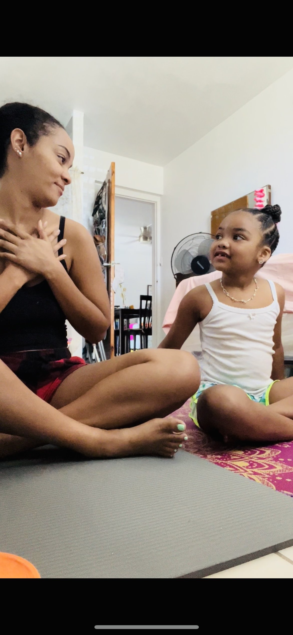 A woman and a young girl sitting on the floor indoors, engaging in a conversation. The woman has her hand on her chest and is smiling, while the girl is looking at her. The background shows a fan, a bed, and a doorway leading to another room.