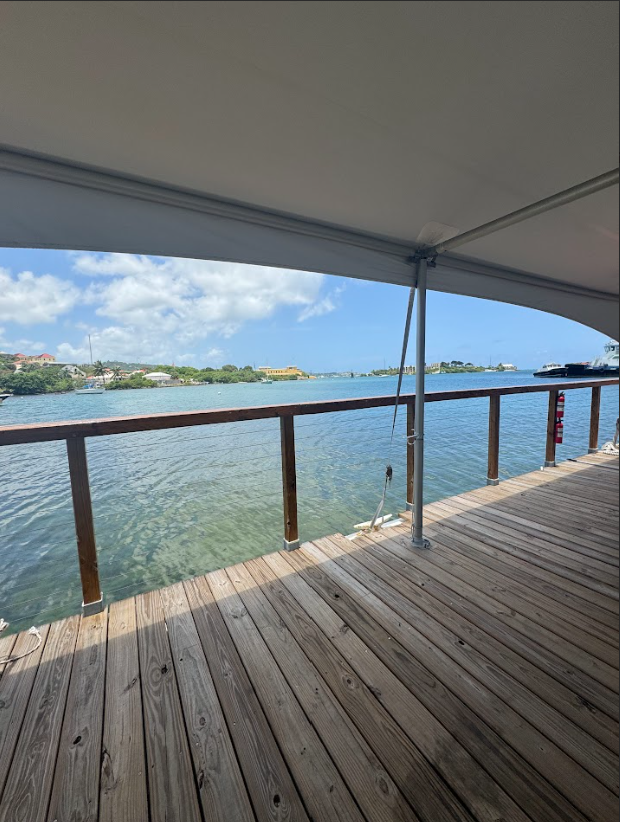 Waterfront deck with wooden floor and railing, sheltered by a canopy, overlooking a calm body of water with buildings and boats in the distance under a partly cloudy sky.